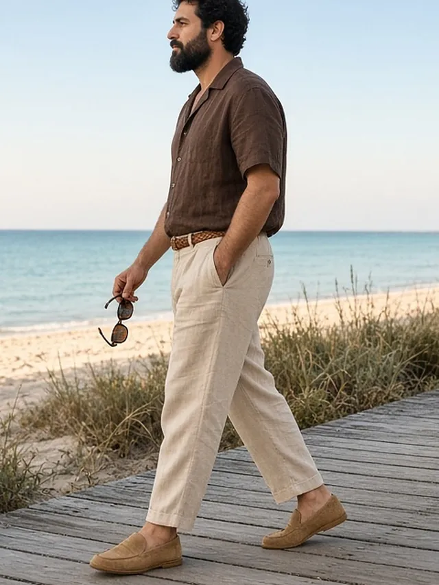 Full-length side-profile photo of a bearded man strolling along a timber beach boardwalk through dune grass, in a chocolate linen camp-collar shirt, ecru linen trousers with a braided belt and tan suede loafers, holding tortoiseshell sunglasses.
